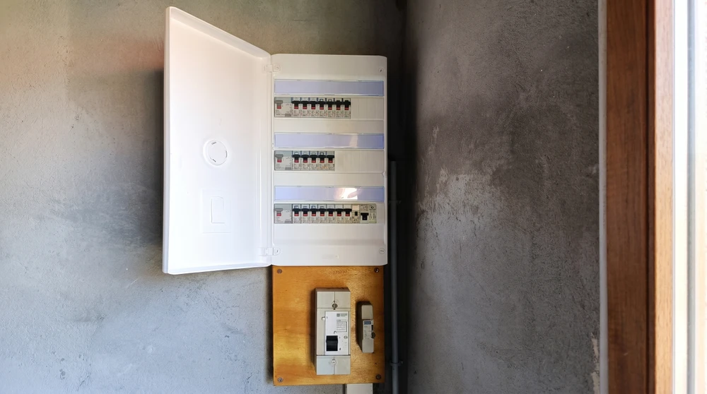 Électricien en salopette bleue et chemise à carreaux consulte un schéma technique devant un tableau électrique ouvert