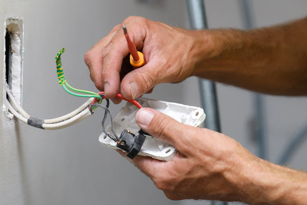 Photographie d'un électricien qualifié en vêtements de travail professionnels intervenant dans un environnement d'entreprise ou d'atelier, effectuant une intervention électrique (installation, câblage ou diagnostic). L'électricien doit être équipé de ses outils professionnels et montrer une approche soignée et professionnelle du chantier. L'arrière-plan doit montrer un espace de travail organisé et respectueux de l'hygiène et sécurité, illustrant ainsi la notion d'"interlocuteur unique" et de "propreté des chantiers" pour les besoins électriques professionnels.