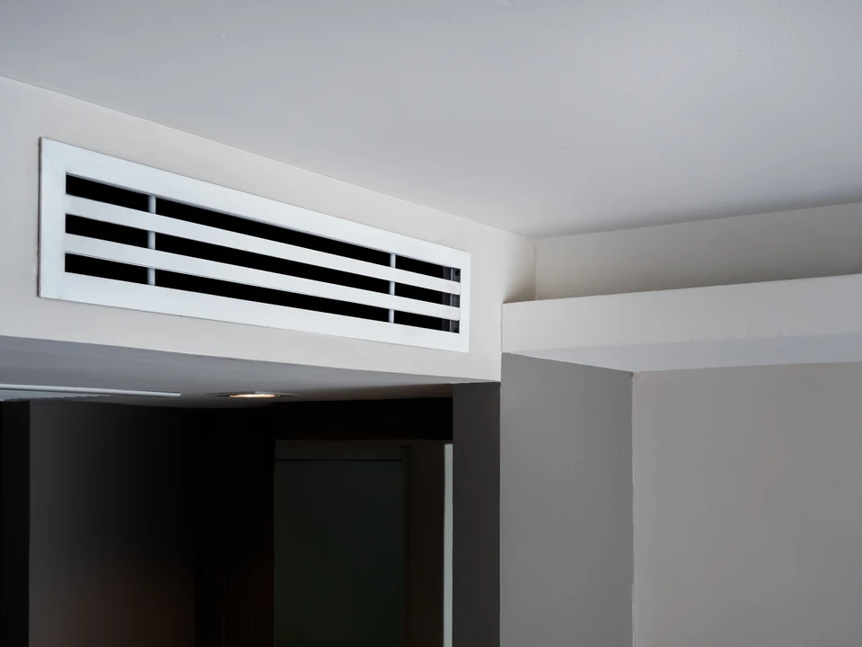 Technicien en combinaison beige avec bandes bleues et rouges installant une grille de ventilation au plafond dans un intérieur résidentiel